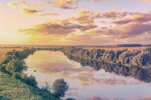 een zonsondergang of zonsopkomst tafereel over- een meer of rivier- met bewolkt luchten reflecterend in de water Aan een zomer avond of ochtend. esthetiek van wijnoogst film. foto