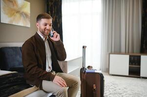 vrolijk zakenman gebruik makend van telefoon zittend in hotel kamer Aan bedrijf reis foto