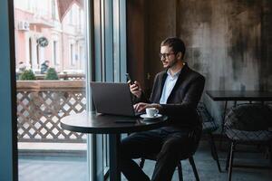 jong zakenman pratend Aan mobiel telefoon terwijl werken Aan laptop in cafe. foto