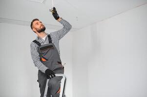 klusjesman of professioneel elektricien in werkkleding installeren licht vlekken, staand Aan de ladder in de wit leven kamer foto