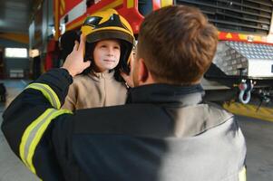 een brandweerman shows zijn werk naar zijn jong zoon. een jongen in een brandweerman helm foto