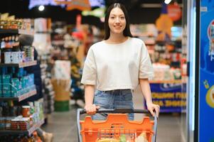 jong glimlachen gelukkig vrouw Jaren 20 in gewoontjes kleren boodschappen doen Bij supermarkt op te slaan met kruidenier kar foto