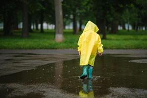 weinig jongen spelen in regenachtig zomer park. kind met paraplu, waterbestendig jas en laarzen jumping in plas en modder in de regen. kind wandelen in zomer regen buitenshuis pret door ieder het weer. gelukkig jeugd. foto