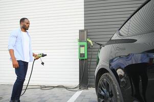 echt Afrikaanse Mens Holding in rekening brengen kabel in Aan hand, staand in de buurt luxe elektrisch auto. foto