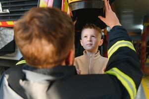 een brandweerman shows zijn werk naar zijn jong zoon. een jongen in een brandweerman helm foto