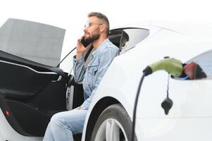 gelukkig Mens gebruik makend van slim telefoon en opladen auto Bij voertuig opladen station foto