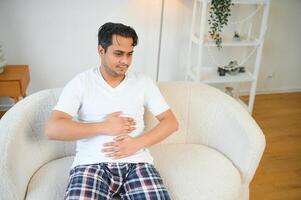 ongelukkig Indisch of Arabisch Mens, zit Aan comfortabel sofa in knus leven kamer, houdt zijn handen Aan zijn maag, grimassen van pijn in zijn maag, lijdt van vergiftiging, spasme, maag problemen. foto