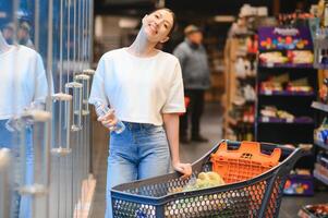 jong tevreden pret vrouw in gewoontjes kleren boodschappen doen Bij supermarkt op te slaan met kruidenier kar foto