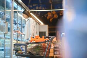 gelukkig jong vrouw op zoek Bij Product Bij kruidenier op te slaan. glimlachen vrouw boodschappen doen in supermarkt foto