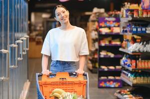 jong glimlachen gelukkig vrouw Jaren 20 in gewoontjes kleren boodschappen doen Bij supermarkt op te slaan met kruidenier kar foto