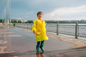 weinig jongen spelen in regenachtig zomer park. kind met paraplu, waterbestendig jas en laarzen jumping in plas en modder in de regen. kind wandelen in zomer regen buitenshuis pret door ieder het weer. gelukkig jeugd. foto