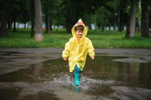 weinig jongen spelen in regenachtig zomer park. kind met paraplu, waterbestendig jas en laarzen jumping in plas en modder in de regen. kind wandelen in zomer regen buitenshuis pret door ieder het weer. gelukkig jeugd. foto