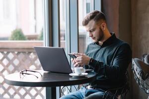 jong zakenman pratend Aan mobiel telefoon terwijl werken Aan laptop in cafe foto