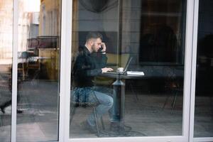 jong zakenman pratend Aan mobiel telefoon terwijl werken Aan laptop in cafe. foto