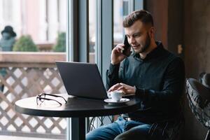 jong zakenman pratend Aan mobiel telefoon terwijl werken Aan laptop in cafe foto