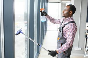 Afrikaanse mannetje professioneel schoonmaak onderhoud arbeider in overall reinigt de ramen en winkel ramen van een op te slaan met speciaal uitrusting foto