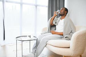 echt jong zwart gebaard mannetje zittend Aan bed in slaapkamer, houden glas van water en tablet Aan wit muur achtergrond foto