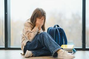 kant visie van verdrietig school- meisje zittend Aan verdieping Bij gang en schuilplaats gezicht door handen en huilen. foto