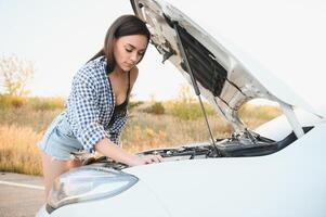 aantrekkelijk slank jong meisje in zomer shorts en overhemd reparaties een gebroken auto. een mooi vrouw staat in de buurt verheven auto kap. foto