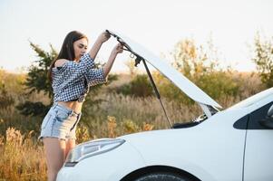 aantrekkelijk slank jong meisje in zomer shorts en overhemd reparaties een gebroken auto. een mooi vrouw staat in de buurt verheven auto kap. foto