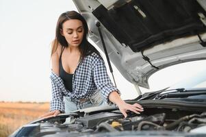 aantrekkelijk slank jong meisje in zomer shorts en overhemd reparaties een gebroken auto. een mooi vrouw staat in de buurt verheven auto kap. foto