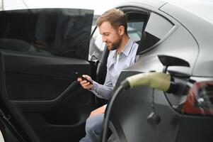 gelukkig Mens gebruik makend van slim telefoon en opladen auto Bij voertuig opladen station foto