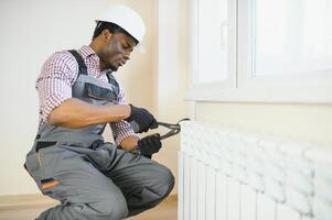 Mens in werkkleding overall gebruik makend van gereedschap terwijl installeren of repareren verwarming radiator in kamer foto