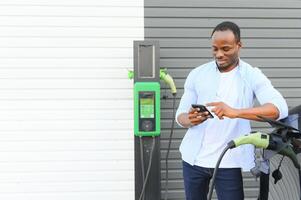 gelukkig jong Mens met mobiel telefoon opladen auto Bij elektrisch voertuig opladen station foto