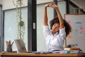 zwart vrouw krijgen fysiek actief van vermoeidheid zittend Bij een bureau. moe van werk, draaien ten gevolge naar pijn, kantoor syndroom van kantoor arbeiders foto