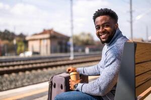gelukkig Mens met koffer geniet drinken koffie terwijl zittend Aan een bank Bij de spoorweg station. foto