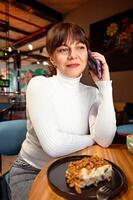 portret van glimlachen vrouw pratend Aan telefoon terwijl zittend in cafe, lunch breken foto