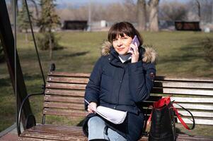 een jong vrouw accountant zit Aan een park bank en leest een rapportage document en oproepen Aan een smartphone, rapportage periode foto
