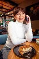 portret van een breed lachend vrouw pratend Aan de telefoon terwijl zittend in een cafe, lunch breken foto