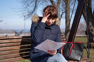 een jong vrouw accountant zit Aan een park bank en leest een accounting document, geklemd haar hoofd, rapportage periode foto