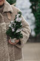 een vrouw in een vacht jas houdt een klein kunstmatig Kerstmis boom in haar handen foto