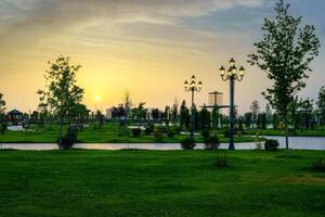 stad park in vroeg zomer of voorjaar met lantaarns, jong groen gazon, bomen en dramatisch bewolkt lucht Aan een zonsondergang of zonsopkomst. foto