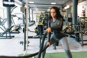 mooi spaans jong vrouw aan het doen sommige crossfit opdrachten met een touw Bij een Sportschool. foto