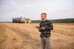 jong aantrekkelijk boer met laptop staand in tarwe veld- met combineren oogstmachine in achtergrond. foto