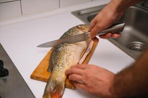 Mens snijdend vers vis in keuken in huis. Mens slachten vis voor koken. foto