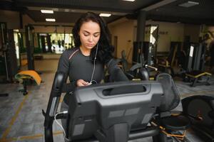 jong vrouw met hoofdtelefoons aan het doen opdrachten Aan schrijfbehoeften fiets in een Sportschool of geschiktheid centrum. jong sportief vrouw in Sportschool luister muziek- van smartphone. Dames aan het doen cardio opdrachten foto