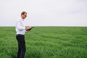 jong agronoom houdt laptop in groen tarwe veld. agribusiness concept. agrarisch ingenieur staand in een tarwe veld- met een tablet in zomer foto