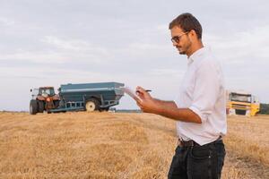 jong aantrekkelijk boer met laptop staand in tarwe veld- met combineren oogstmachine in achtergrond. foto