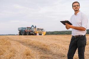 gelukkig jong boer ingenieur met notitieboekje staand Aan tarwe veld- terwijl combineren oogstmachine werken in achtergrond. foto