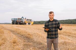 jong agronoom Mens staand Aan tarwe veld- controle kwaliteit terwijl combineren oogstmachine werken foto