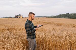 jong aantrekkelijk boer met laptop staand in tarwe veld- met combineren oogstmachine in achtergrond. foto