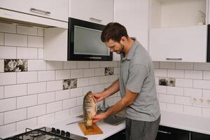 Mens snijdend vers vis in keuken in huis. Mens slachten vis voor koken. foto