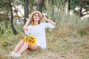 buitenshuis schot van aangenaam op zoek vrouw met gebruind gezond huid, gekleed in wit jurk en zomer hoed, poses in park met zelfverzekerd tevreden uitdrukking, houdt recreatie. mooi jong vrouw. foto