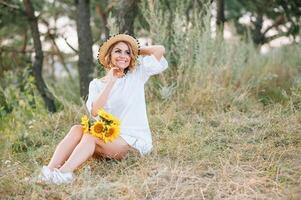 buitenshuis schot van aangenaam op zoek vrouw met gebruind gezond huid, gekleed in wit jurk en zomer hoed, poses in park met zelfverzekerd tevreden uitdrukking, houdt recreatie. mooi jong vrouw. foto