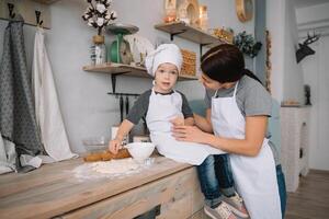 jong gelukkig mam en haar baby koken koekjes Bij huis in de keuken. Kerstmis eigengemaakt ontbijtkoek. schattig jongen met moeder in wit uniform en hoed gekookt chocola koekjes. foto