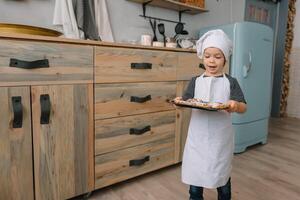 jong jongen schattig Aan de keuken koken chef in wit uniform en hoed in de buurt tafel. Kerstmis eigengemaakt ontbijtkoek. de jongen gekookt de chocola koekjes foto